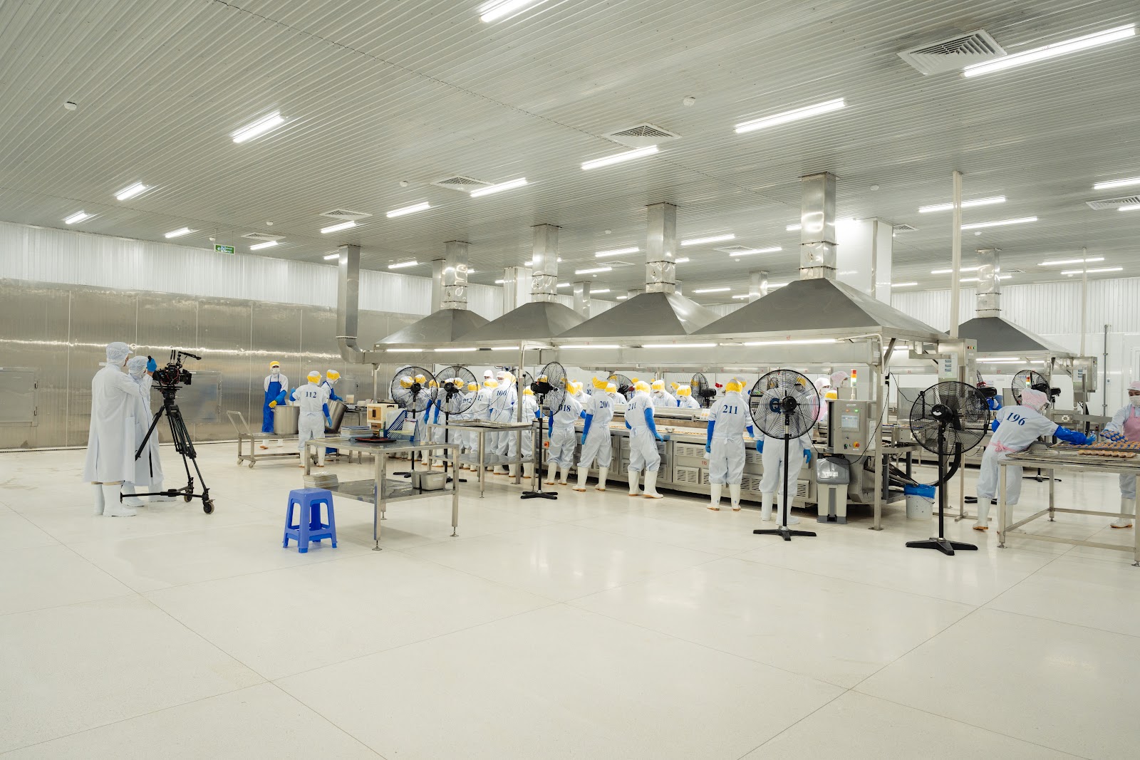 CINEFY crew setting up a camera on a tripod at the edge of a food processing hall while workers in protective gear work at steel tables in the background